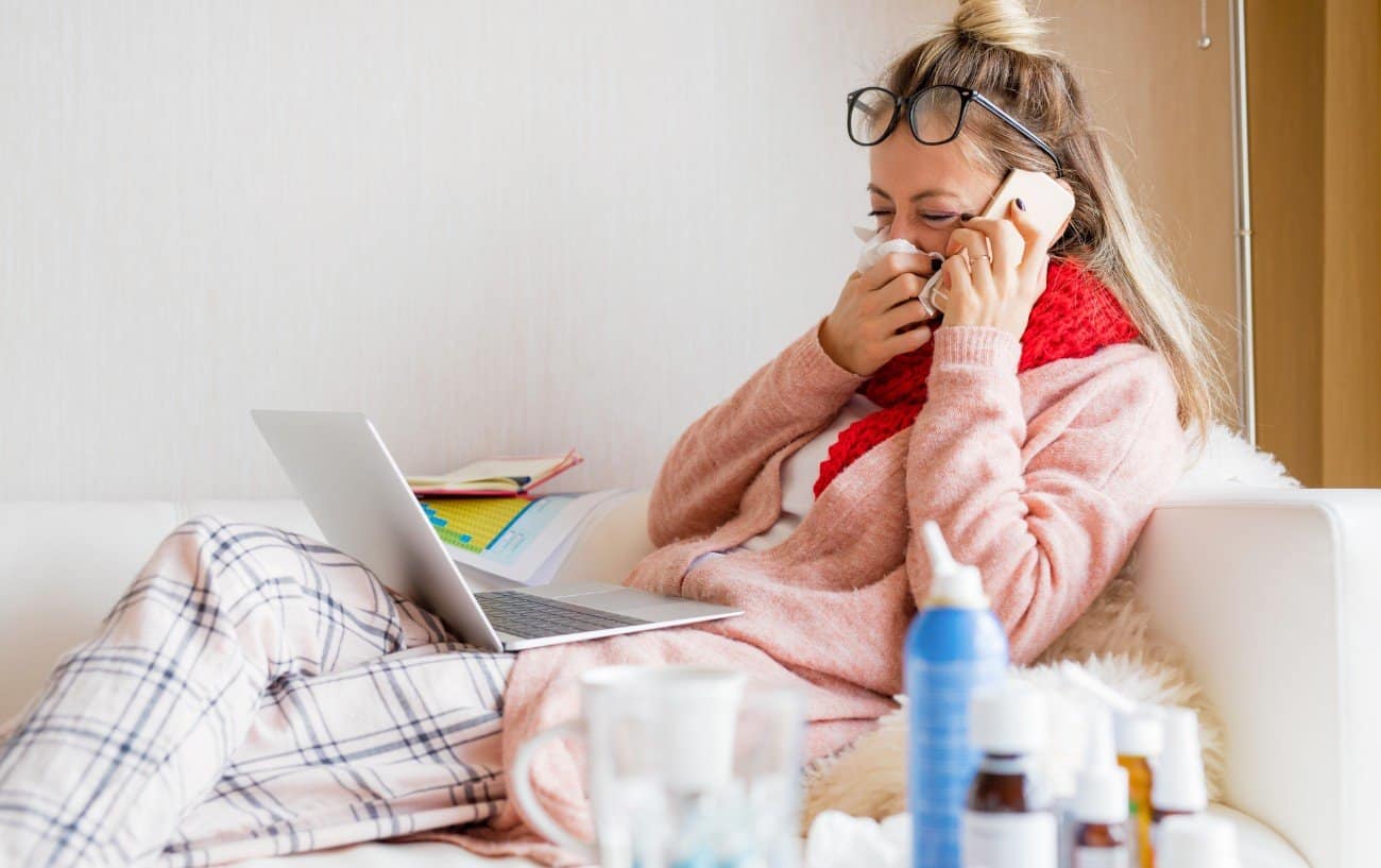 Woman sick on a couch blowing her nose looking at the computer.