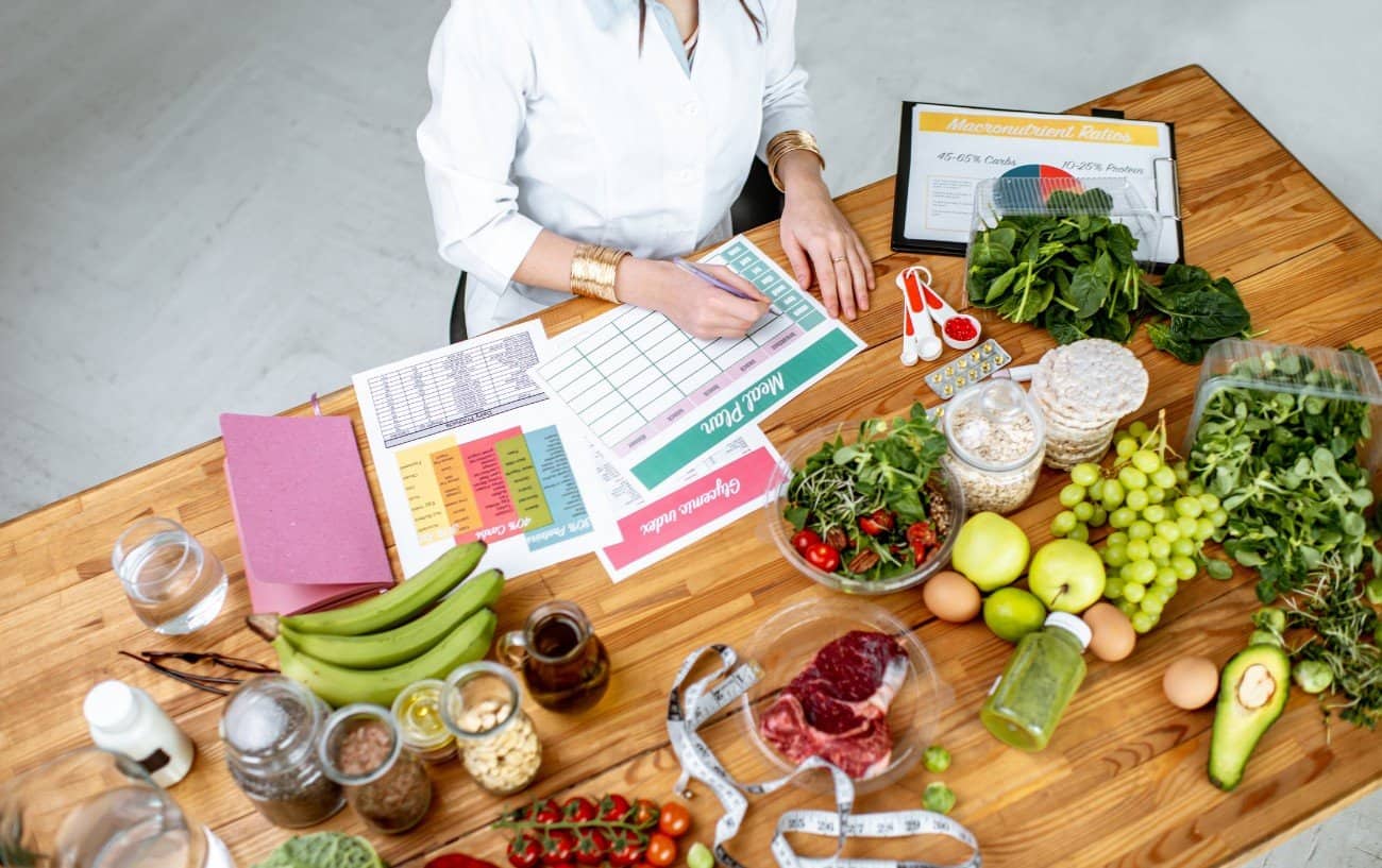 Una nutricionista sentada en una mesa llena de diferentes alimentos saludables, preparando un plan de comidas.