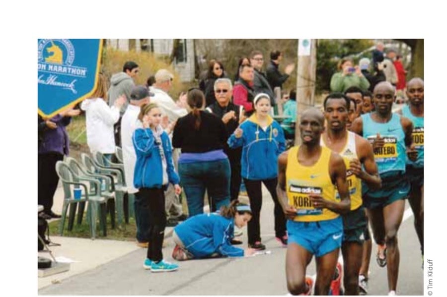 Framingham High School classmates Hannah Greenberg (delivering data by phone), Isabelle Tambascio (kneeling, recording splits), and Grace Seta (signaling) work together as part of the Spotters Network during the 2013 Boston Marathon. They are carefully watching Wesley Korir of Kenya lead Markos Geneti of Ethiopia, Levy Matebo of Kenya, and Micah Kogo of Kenya.