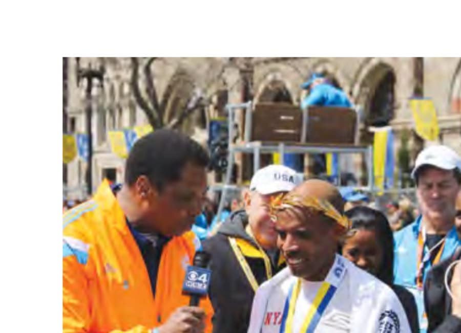 At the 2014 Boston Marathon, champion Meb Keflezighi of the United States wears his gold-dipped wreath while being interviewed by WBZ-TV 4 Boston sportscaster Steve Burton, as Meb's coach (Bob Larsen), wife (Yordanos Asegdom), and brother (Merhawi), from left, look on proudly behind him.
