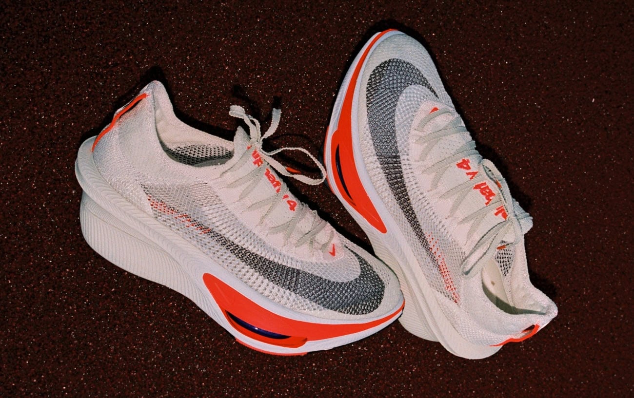 A pair of white Nike Alphafly 4 racing shoes with orange and grey detailing, photographed on a dark red track surface.